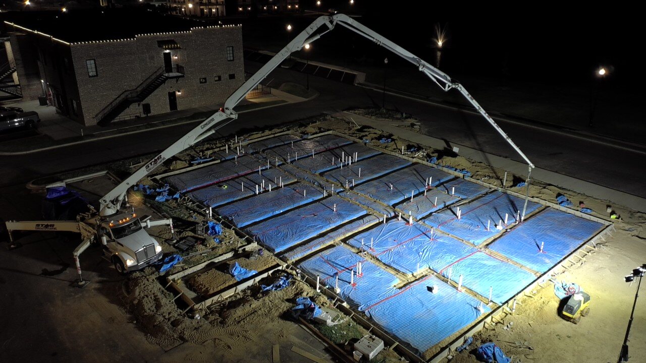 Nighttime aerial view of a concrete pump truck placing a large foundation slab pour for a new ground-up commercial build